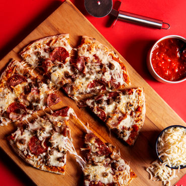 A Meat Lovers pizza on a wooden cutting board with cheese and sauce beside it