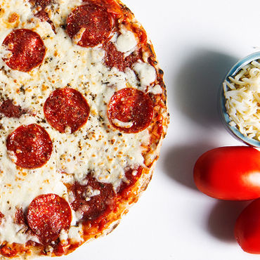 A close up picture of a Pepperoni pizza with a bowl of cheese and two tomatoes beside it.