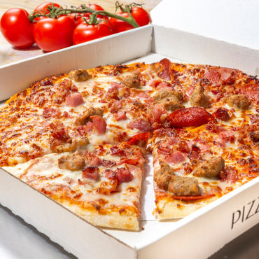 A meat lovers pizza in a box with tomatoes behind it