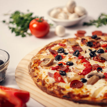 A Vegetarian pizza on a wooden cutting board with olives, garlic, and tomatoes beside it