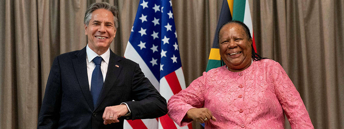 Antony Blinken and Naledi Pandor facing the camera side-by-side greeting each other with an elbow bump. The US and South African flags are behind