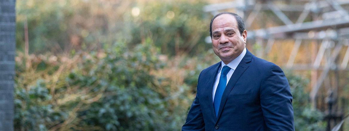 Egyptian President Abdel Fattah El-Sisi walking in front of vegetation.