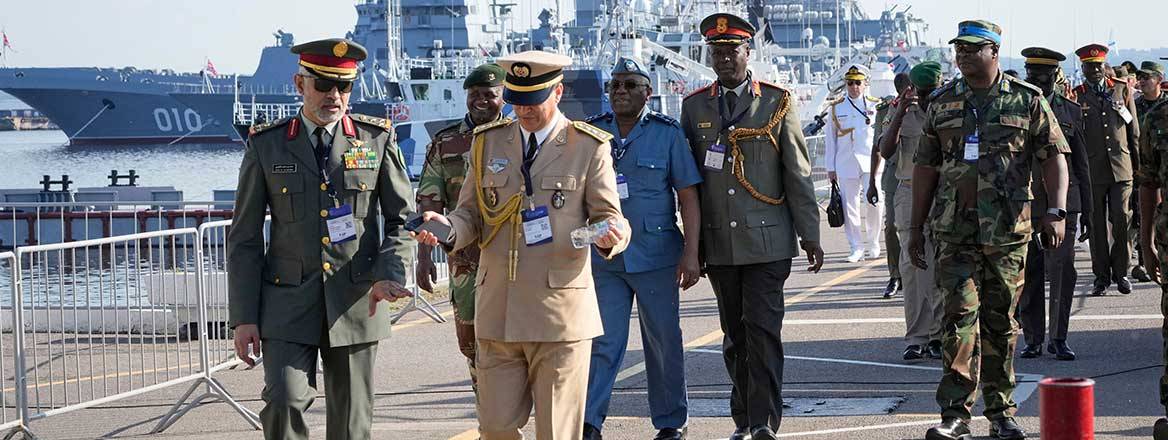 Group of military personnel walking on a paved path along a shoreline with a military ship in the background