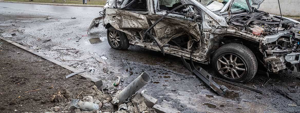 A destroyed car on a road with the remains of a munition next to it