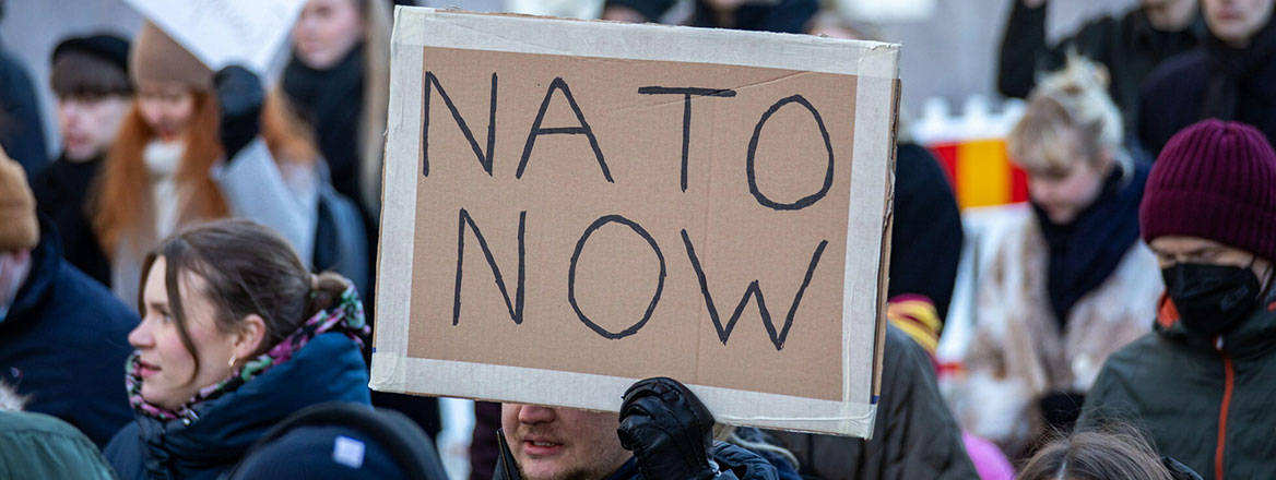 A cardboard sign with handwritten 'NATO NOW' written on it with a large crowd in the background