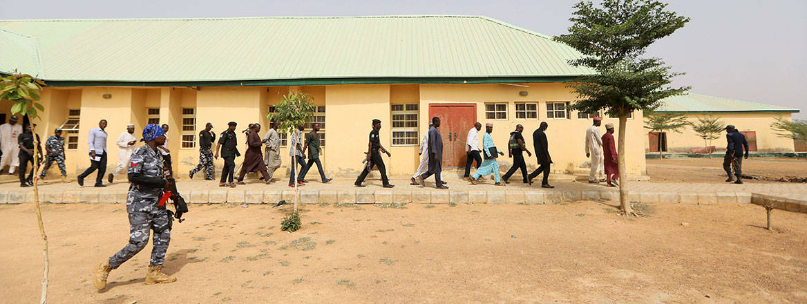 A pale-coloured single-storey building with several people, some in uniforms, walking in front of it. The ground is sparsely vegetated and dry.