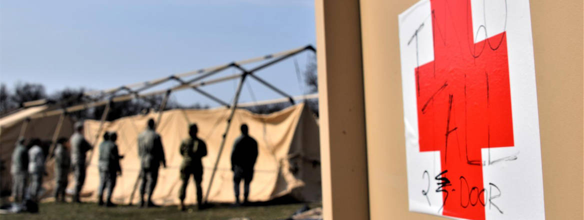 Airmen put up tent at Role 2 (field hospital) expeditionary medical support facilities at Cincu Military Base, Romania, April 4, 2019. A close up of a red cross on a box is shown to the right of the image