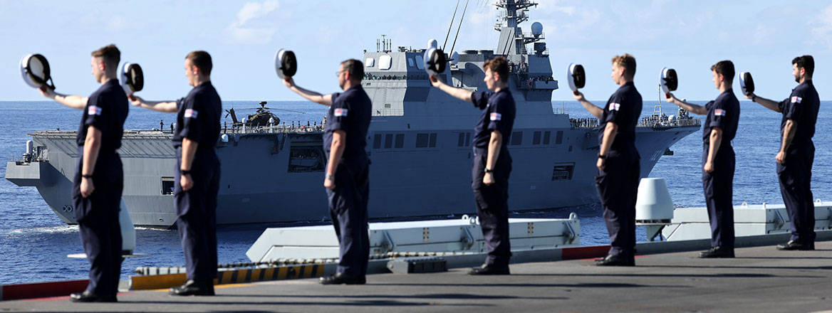 Several uniformed naval personnel standing in line on the edge of a ship holding caps in front in the direction of a passing ship