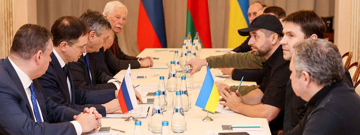 Russian negotiators (left) sitting on a table with a white cloth opposite Ukrainian negotiators (right)