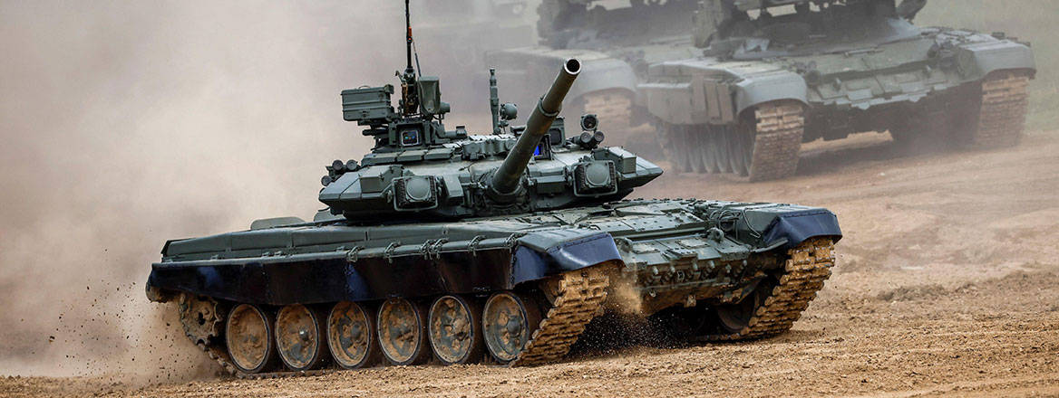 A Russian T-80 tank driving on a rough, brown-coloured surface with other military vehicles in the background.