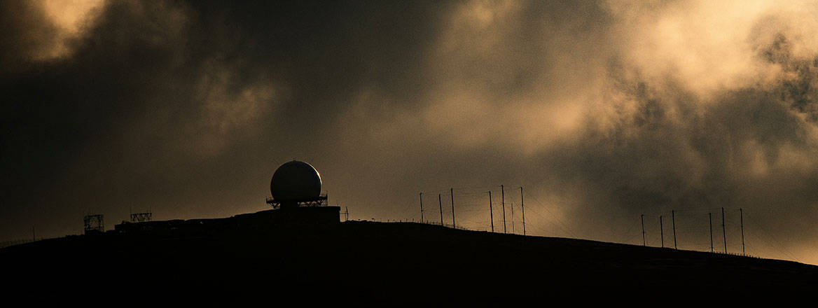 A long distance shot of a radar station sitting on top of a hill. Sky is cloudy, dark but with some light creating a shadow effect