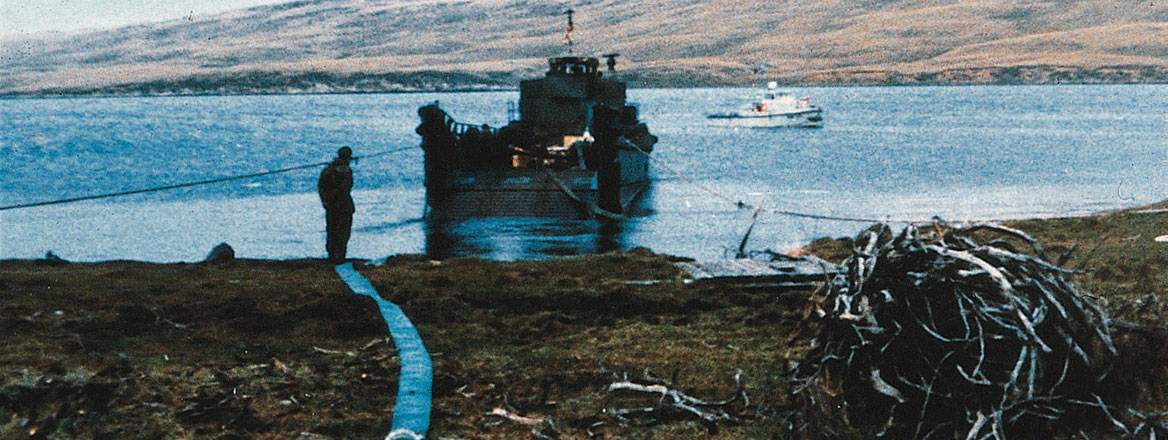 A small ship ashore with a person standing next to it with a fuel pipeline on the ground