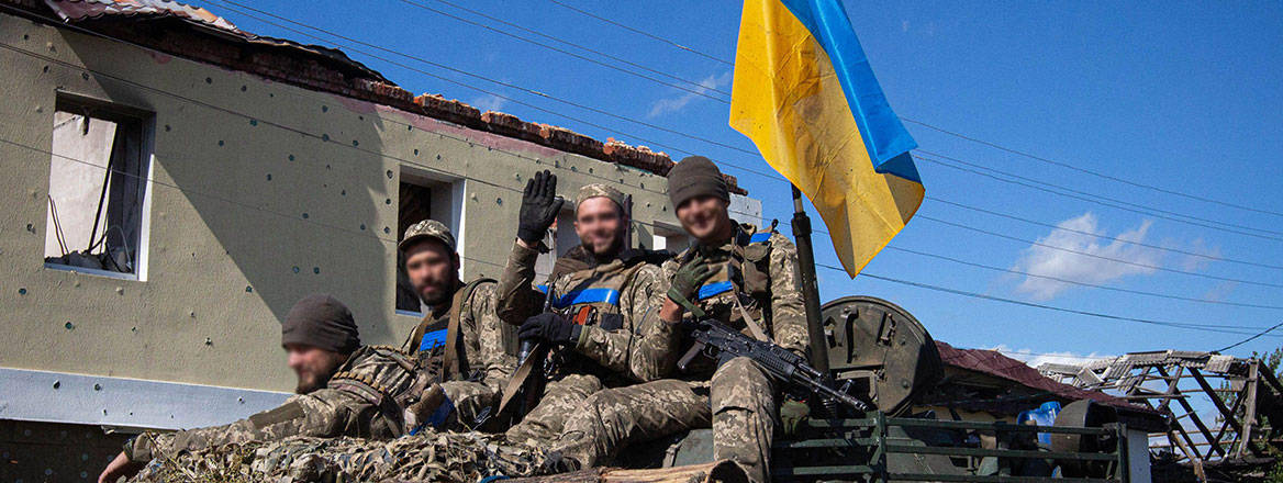 Ukrainian personnel on top of a vehicle facing the camera with a Ukrainian flag slightly behind them