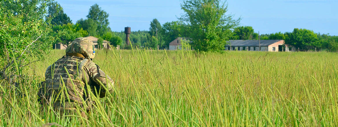 A Ukrainian soldier with back facing the camera blends into long green grass with a few small buildings in the distance