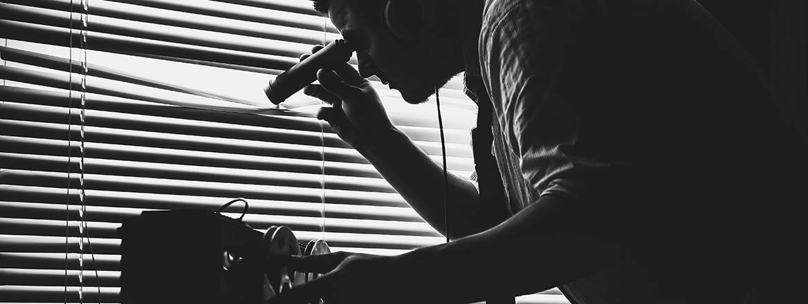 Stock image of a man in office clothes wearing can-headphones looking though the blinds of a window with a pair of binoculars while he a tape recorder operates in the foreground.