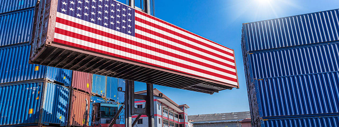 Stock image of a cargo container being carried by a forklift truck through a stak of other  containers. The  elevated container  is illustrated here with an American flag on the broadest side. 