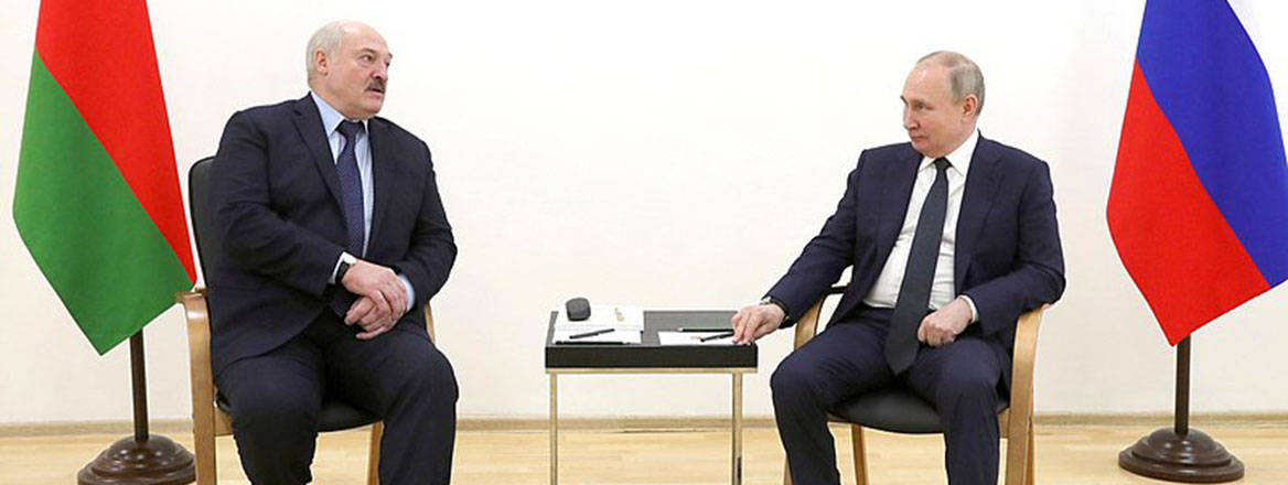 Alexander Lukashenko (left) and Vladimir Putin (right) sit next to each other with a small table in between and national flags behind them