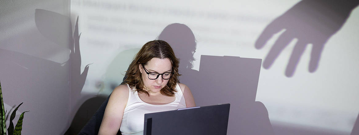 A stock image of a researcher working on a laptop while an unseen hand casts a shadow that reaches sinisterly towards her computer.