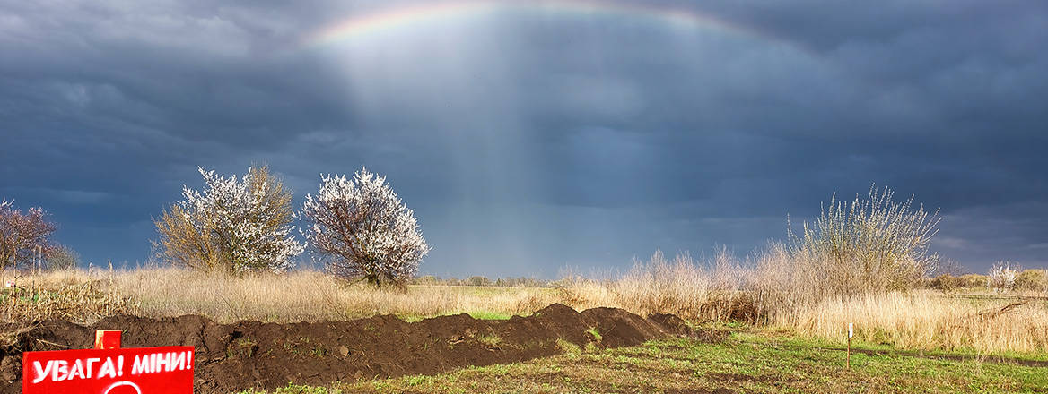 A rainbow forms against a cloudy sky above a country field marked with red sign warning in Ukrainian of landmines.