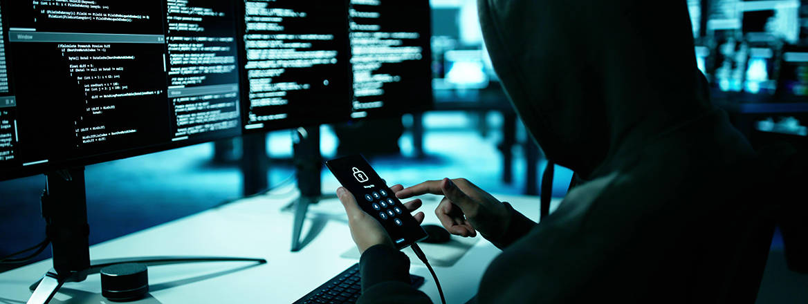 Stock image of a hooded figure with a phone at a computer terminal.