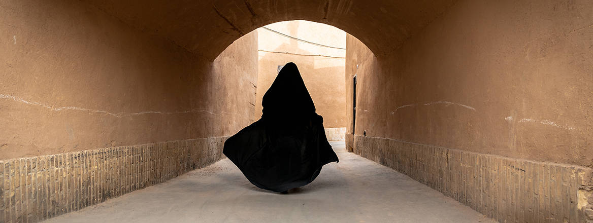 In Iran, an unidentified woman, dressed in a black hijab, walks through a narrow street of old Yazd city, reaching the threshold of a tunnel into broad daylight.