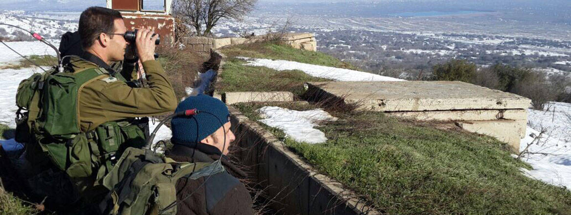 Soldiers standing on top of partially snow-covered high ground looking into the distance, one through binoculars