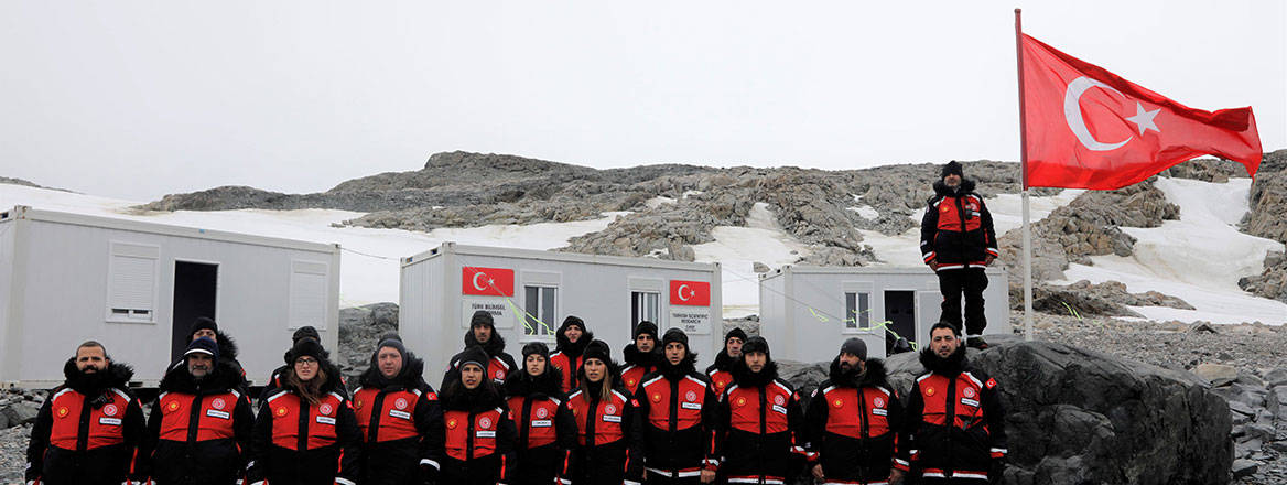 A team from Turkey posing with the Turkish flag in front of some small white structures with a rocky and icy backdrop further behind.