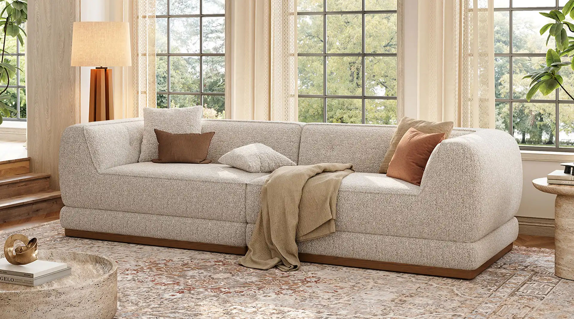 Light gray curved sofa with rounded arms, accent pillows, and a draped throw in a bright living room with large windows.