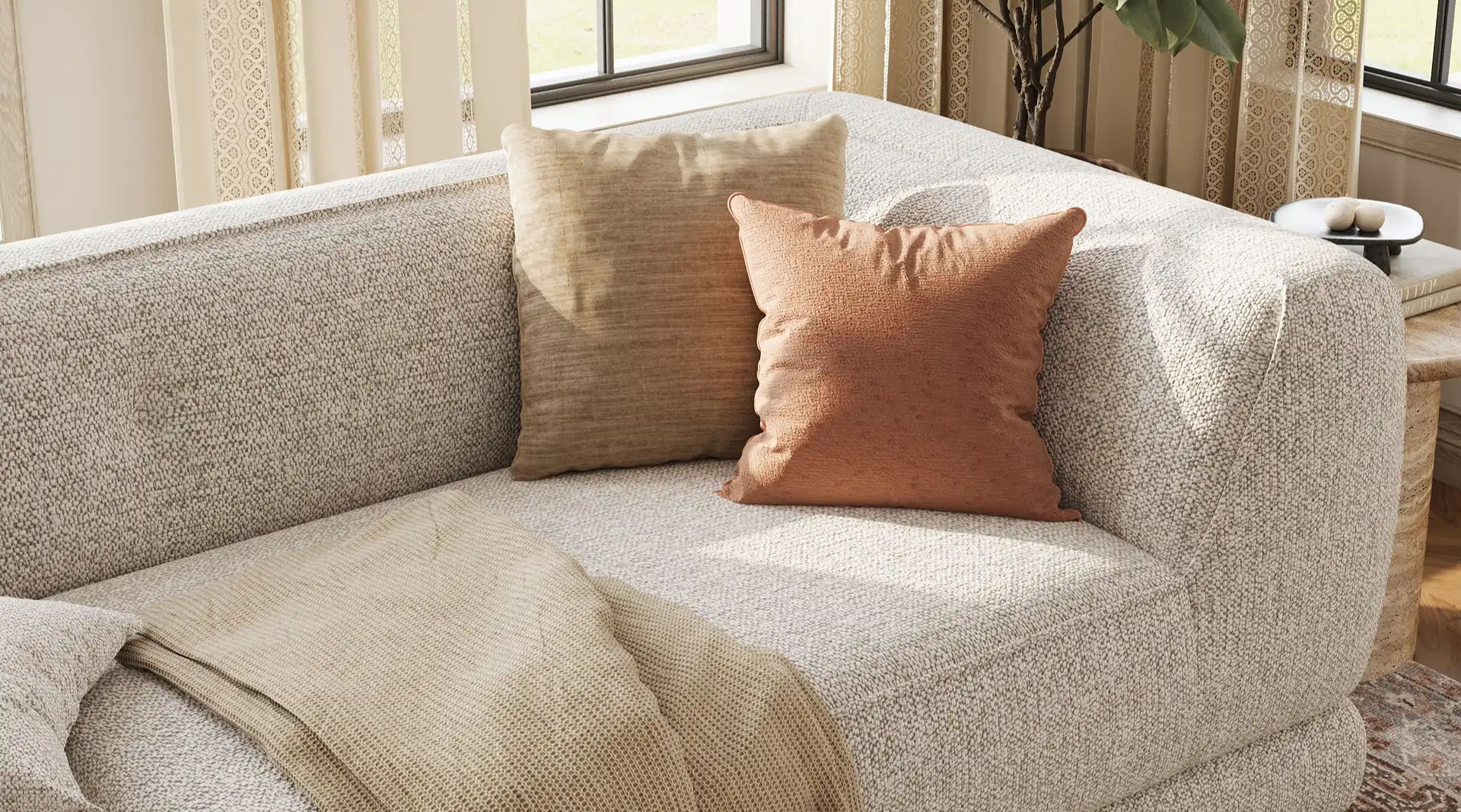 Close-up of a light gray textured sofa with wide rounded arms, neutral accent pillows, and a beige throw in a sunlit living room.