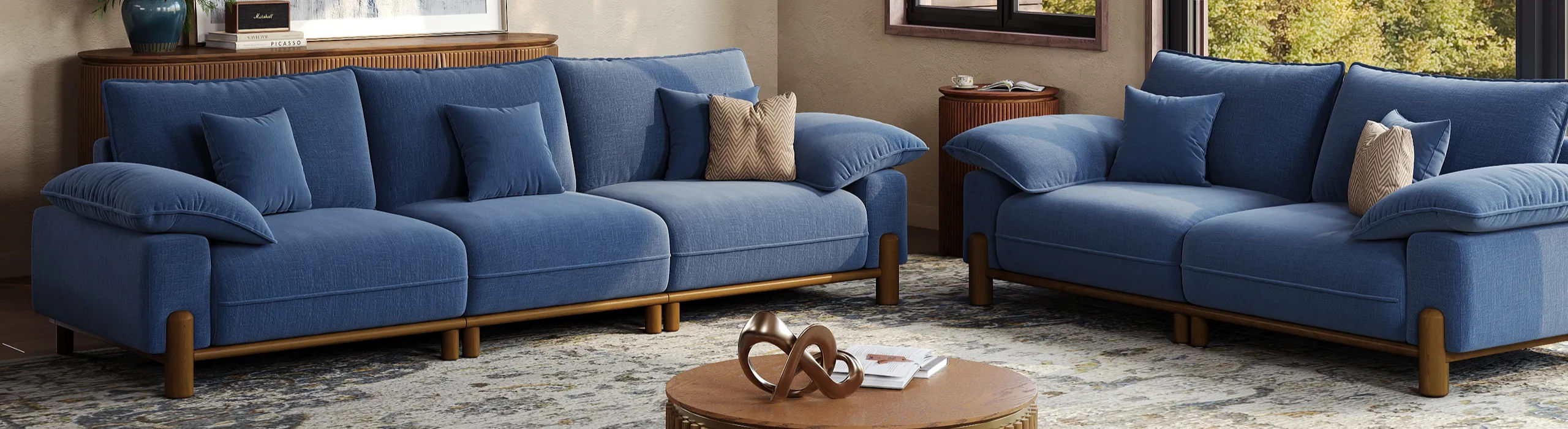Two plush navy blue sofas with soft throw pillows are arranged facing each other in a bright, sunlit living room with high ceilings.