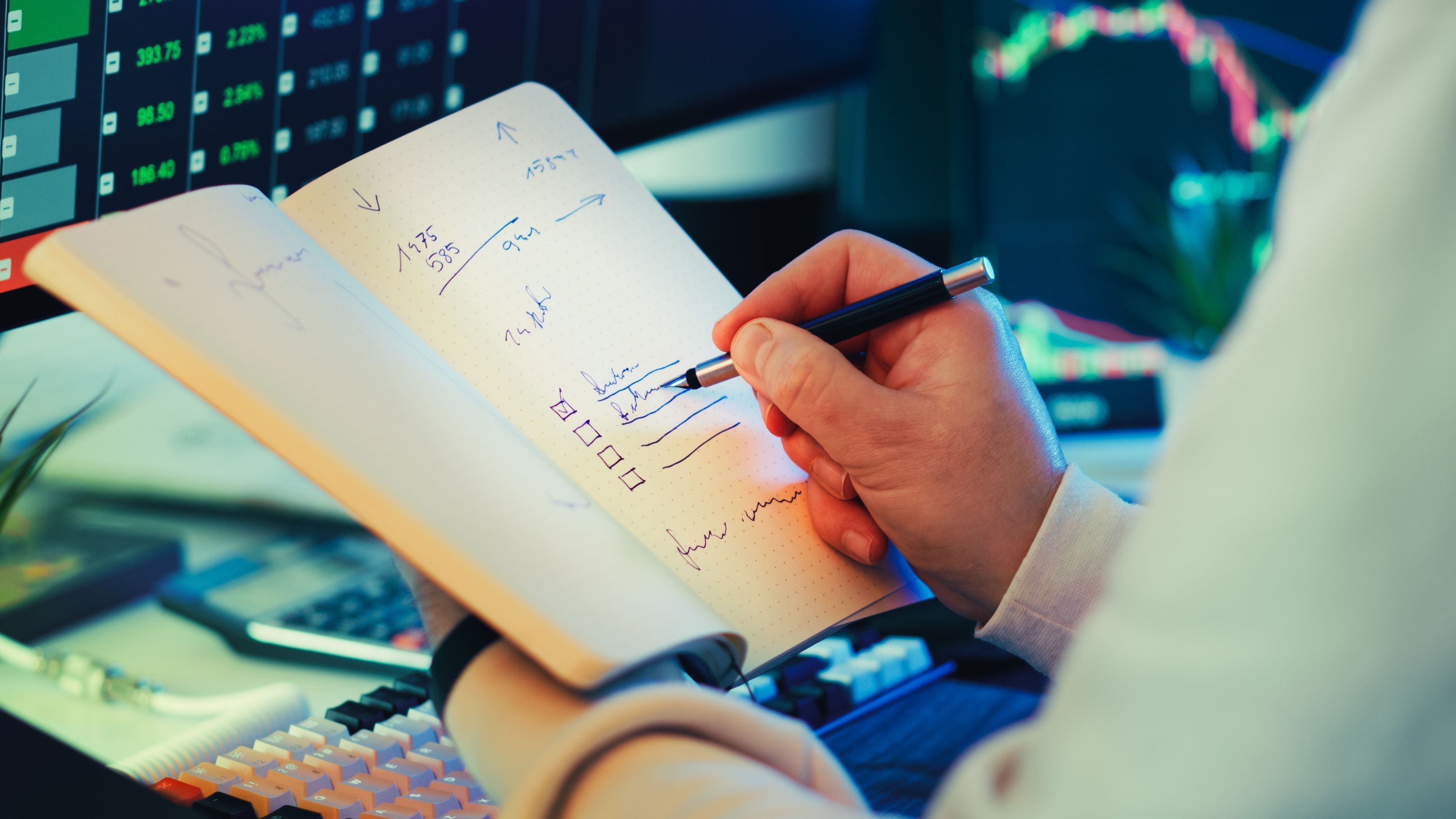 Financial trader taking notes and planning stock investment strategies in front of multi-monitor trading charts.