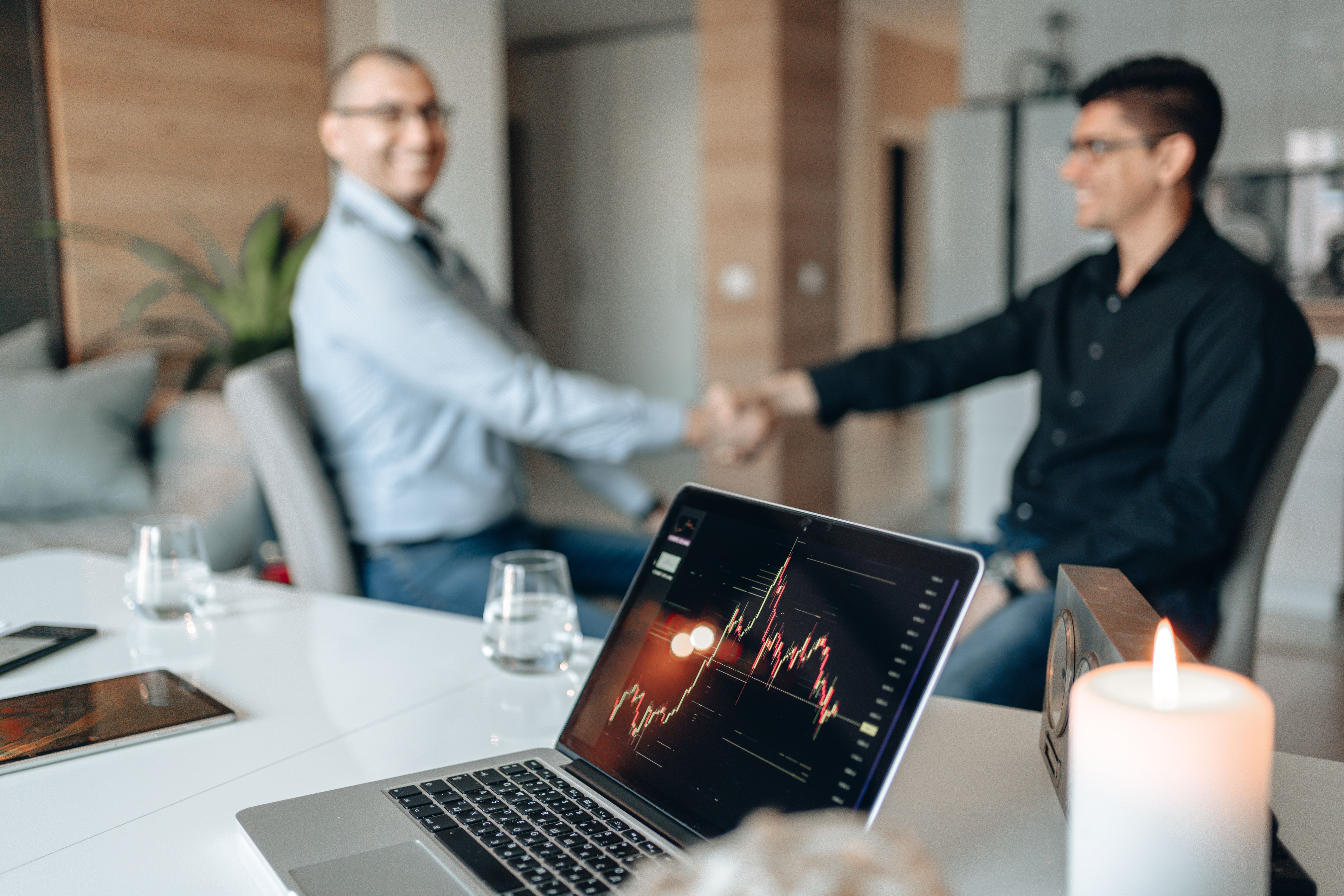 Two businessmen shaking hands in an office with a laptop displaying financial stock charts in the foreground.