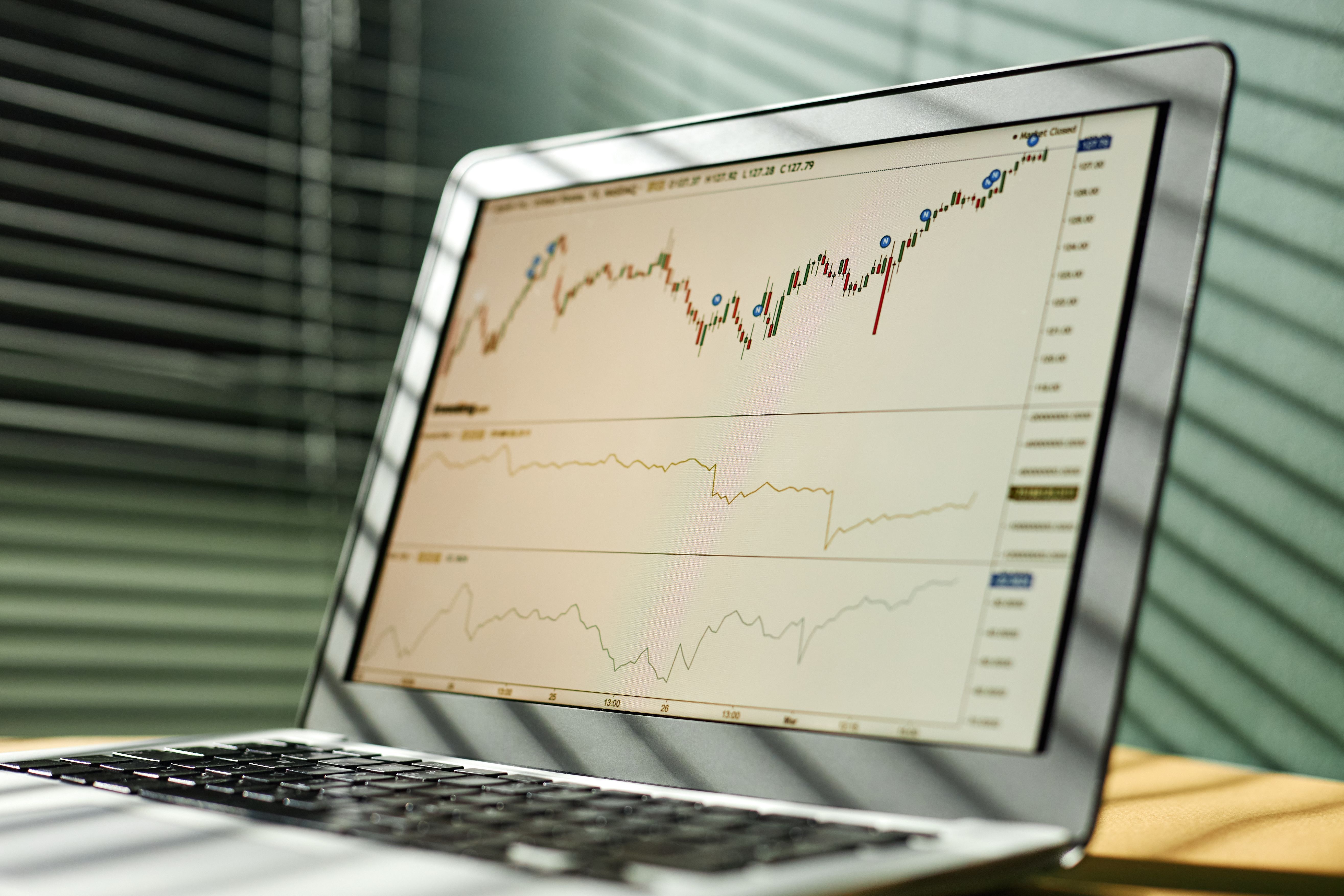 Laptop displaying live stock market candlestick graphs on a glass table next to a notebook