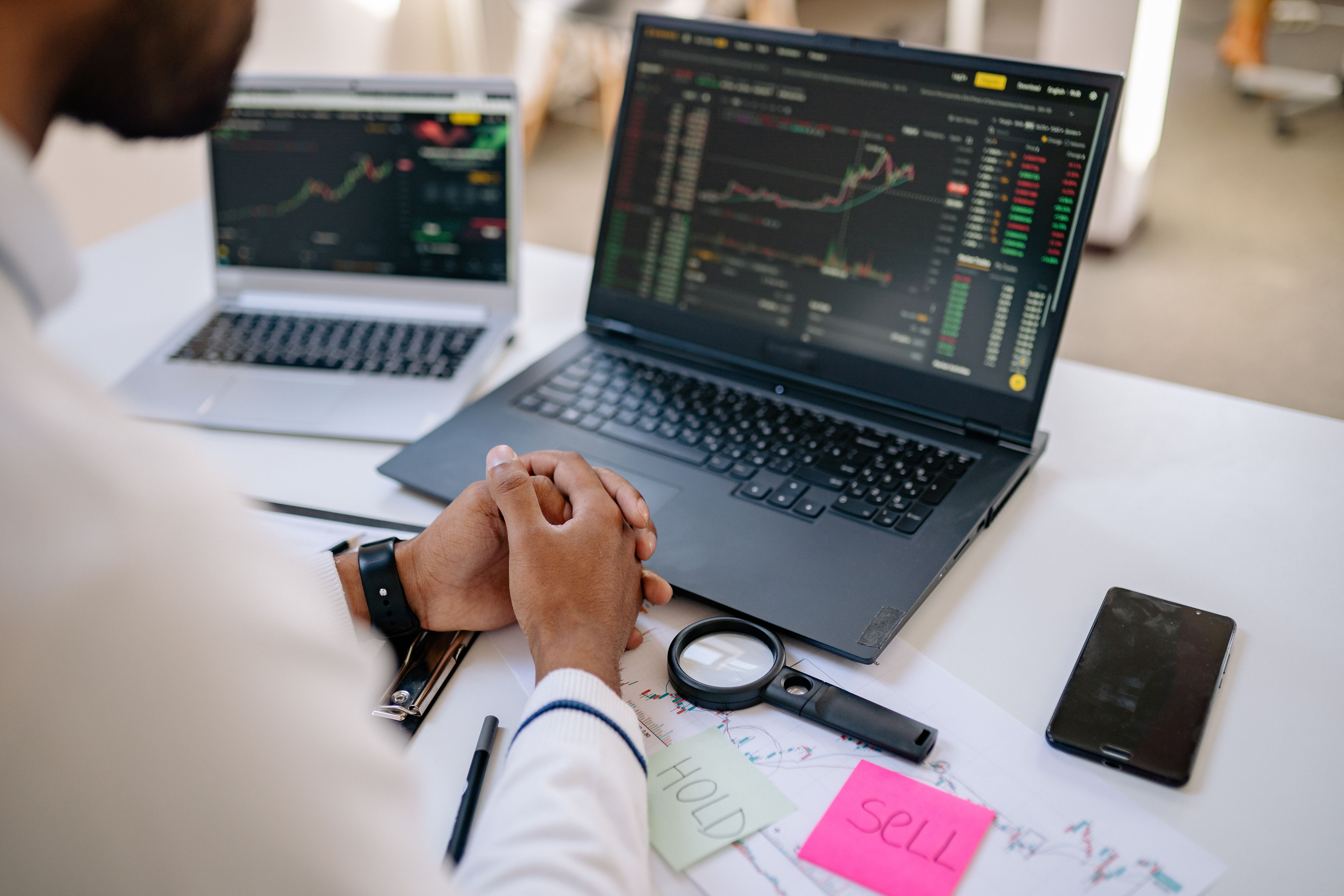Financial trader at desk with laptops showing stock charts, sticky notes reading Hold and Sell, and a magnifying glass.