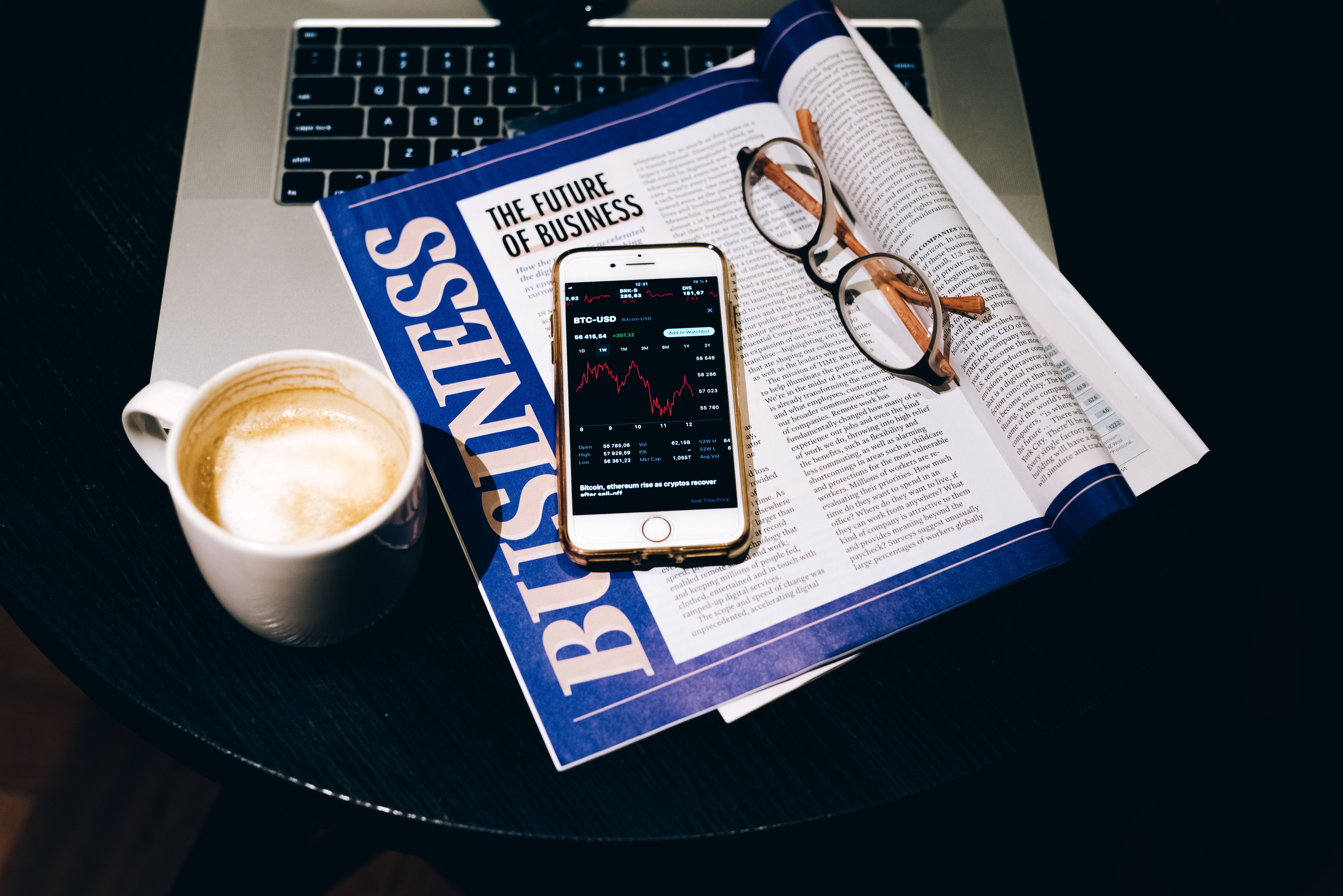 Top view workspace with a smartphone displaying bitcoin trading data on top of a business newspaper, coffee cup, and reading glasses