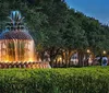 The image shows a large pineapple-shaped fountain with water cascading down its tiers surrounded by lush greenery and trees in a park-like setting during dusk or early evening