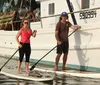 Two people are standing on paddleboards next to a moored fishing boat in calm waters each holding a paddle