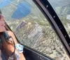 A person wearing sunglasses and a headset is looking out the window of a helicopter at a scenic landscape below