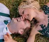 A man and a woman likely a couple are lying on the grass sharing a joyful moment with the woman on top both dressed in formal attire with the man wearing a suit and the woman in a dress with a floral design