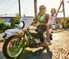 Two individuals are joyfully riding on a classic motorcycle with the passenger waving on a sunny day down an urban street lined with cobblestones and industrial buildings