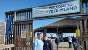 Two individuals are smiling in front of a Welcome sign at Tybee Island, which is known as Savannah's Beach, indicating a popular tourist destination.