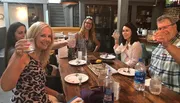 A group of five people is cheerfully toasting with drinks at a dining table scattered with finished plates and glasses.