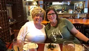 Two women are smiling at the camera while seated at a restaurant table with drinks and a plate of food in front of them.