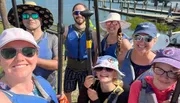 A group of eight smiling people equipped with paddles and wearing life jackets and sun hats appears ready for a kayaking adventure by the water.