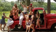 A group of cheerful women poses with a vintage red pickup truck in a sunny, outdoor location, likely celebrating a festive occasion.