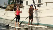 Two people are standing on paddleboards next to a moored fishing boat in calm waters, each holding a paddle.