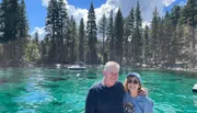 A smiling couple poses for a photo with a backdrop of crystal-clear turquoise water and a lush pine forest under a blue sky.