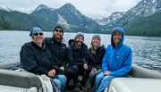 A group of five people, wearing warm clothes and smiles, are seated on a boat with a scenic backdrop of a mountainous, forested area and overcast skies.