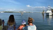 Three individuals are observing a waterfront scene with boats and a bridge in the distance from a dock or a pier.