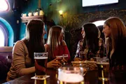 A group of people is enjoying a conversation over pints of beer in a cozy pub with ornate wallpaper and a blue tinted window.